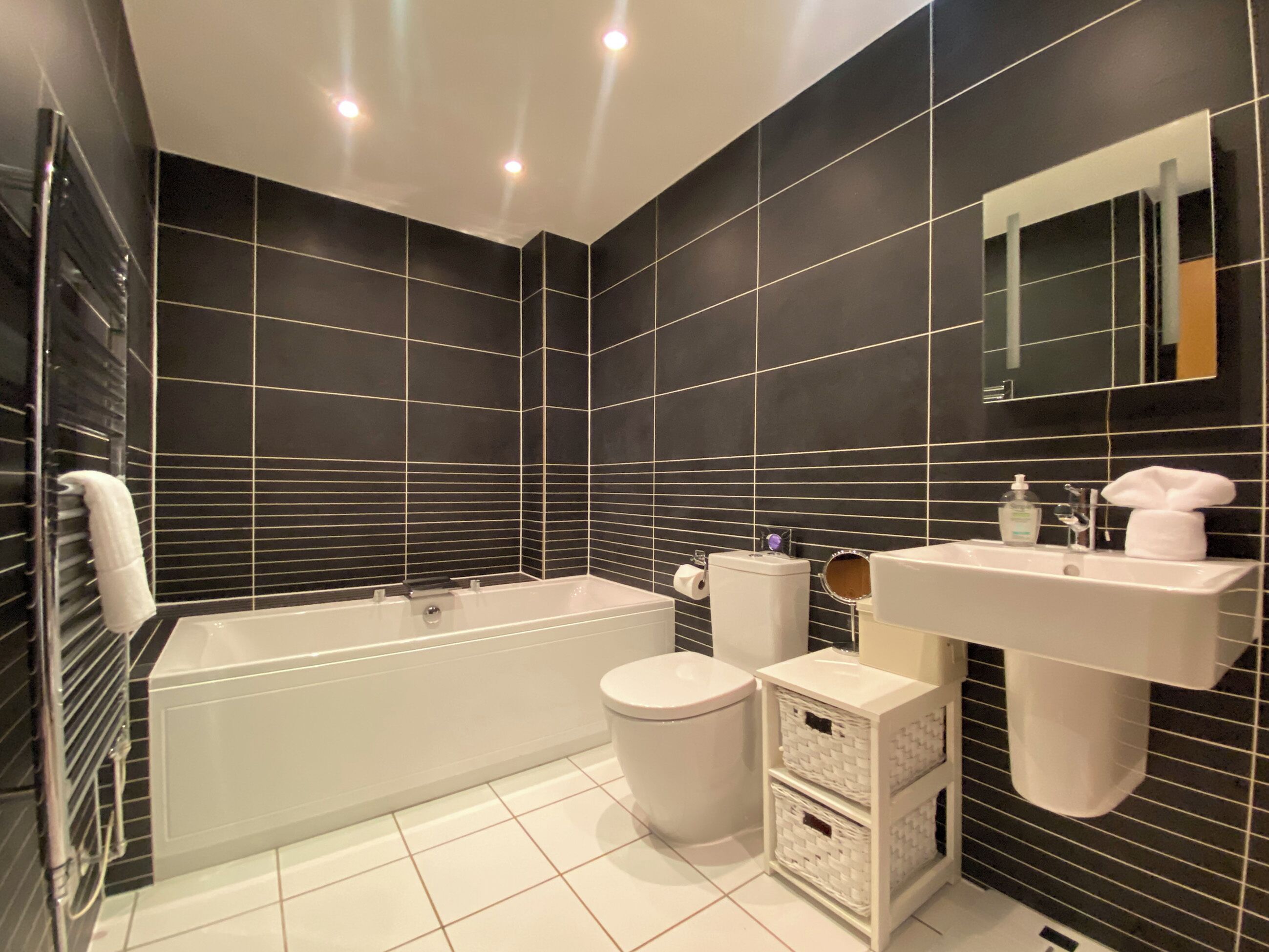 Un baño moderno con bañera, inodoro y lavamanos, con paredes de azulejos negros y sanitarios blancos.