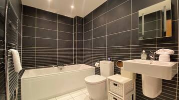 Un baño moderno con bañera, inodoro y lavamanos, con paredes de azulejos negros y sanitarios blancos.