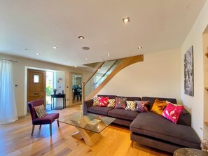 Un salon moderne avec une table basse en verre, un canapé marron foncé orné de coussins à motifs, une chaise en bois et un escalier menant à un niveau supérieur.