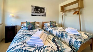 A bedroom with two beds, a nightstand, a lamp, a wall-mounted shelf, and a picture.