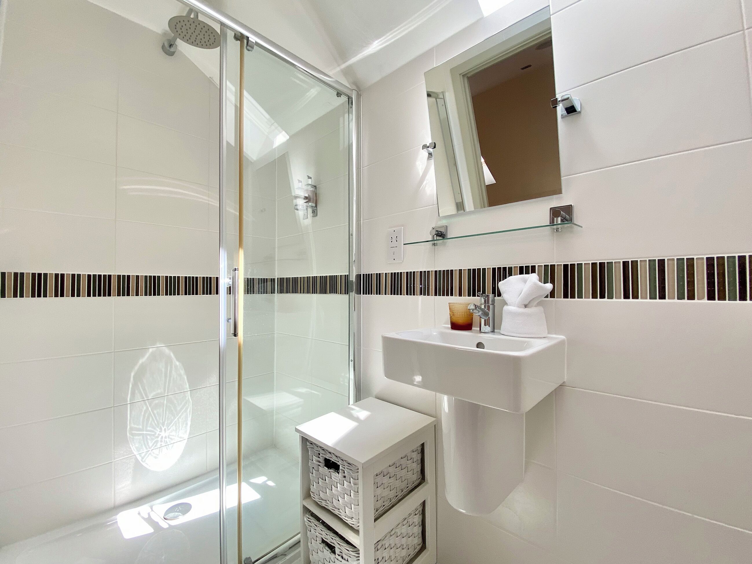 Une salle de bain moderne avec une douche à l’italienne en verre, un lavabo blanc avec un miroir au-dessus et un pare-chocs en carreaux à rayures.