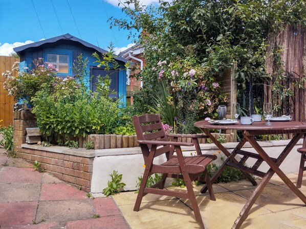 A patio with a table and chairs, surrounded by lush greenery and a blue building.