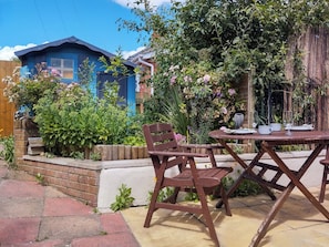 A patio with a table and chairs, surrounded by lush greenery and a blue building.