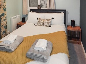 A neatly made bed with a brown throw blanket, two rolled towels, and a decorative pillow with animal prints. There are two bedside tables with lamps, a window with curtains, and a radiator.