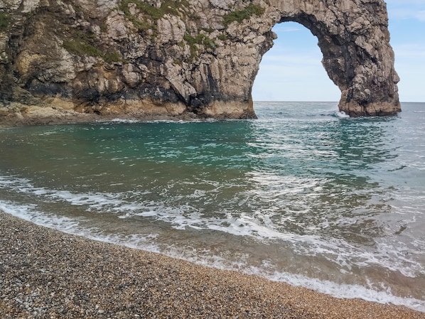 A coastal scene with a large rock arch formation, clear turquoise water, and a pebbled beach.