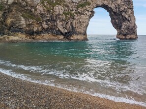 A coastal scene with a large rock arch formation, clear turquoise water, and a pebbled beach.