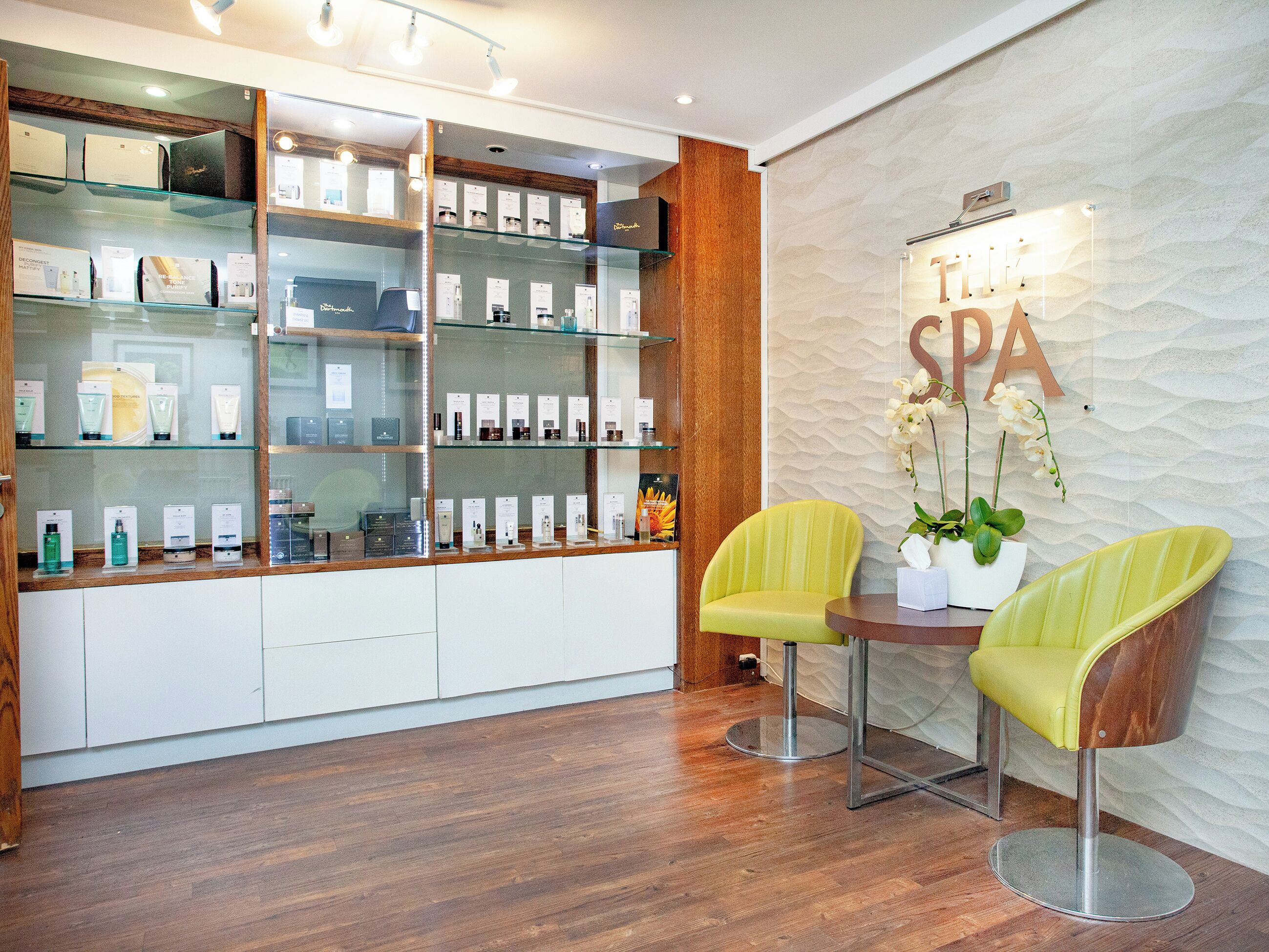 Een receptiegedeelte van een spa met een houten balie, glazen vitrines en twee gele stoelen.