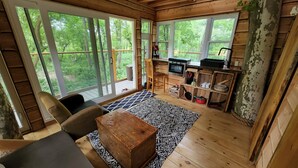 A rustic cabin interior with a wooden floor, a sofa, a coffee table, a kitchen area with a stove, and large windows offering a view of the forest. - One of a kind Treehouse on the Genesee River (BELFAST)