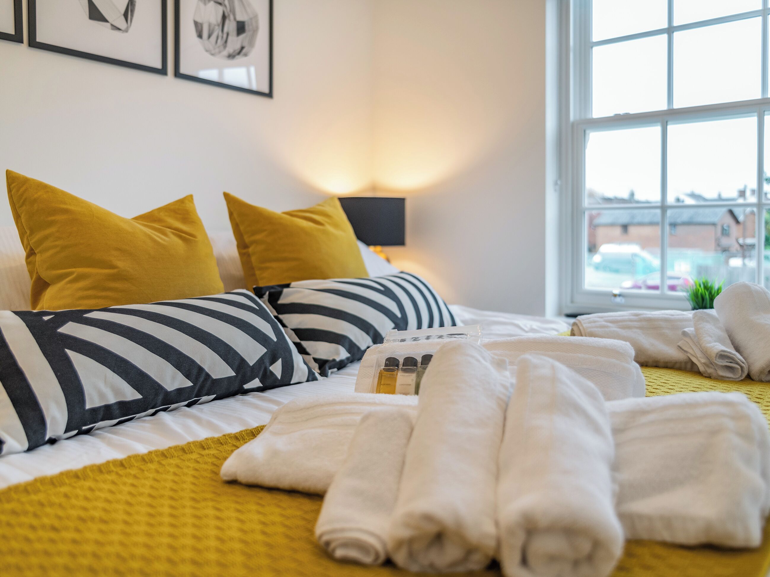 Una cama bien hecha con almohadas a rayas, una colcha amarilla y toallas enrolladas sobre un edredón amarillo.