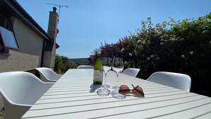 A table with a bottle and glasses, surrounded by white chairs, in a sunny outdoor setting.