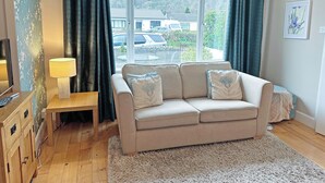 A living room with a beige sofa, a wooden side table, a lamp, and a view of a residential area through large windows. - 2 bedroom accommodation in Lamlash, Isle of Arran (Lamlash, Isle of Arran)