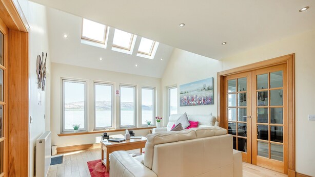 A spacious living room with a large white sofa, a wooden floor, and a view of the sea through multiple windows. - 3 bedroom accommodation in Port Lamont near Dunoon (Port Lamont near Dunoon)