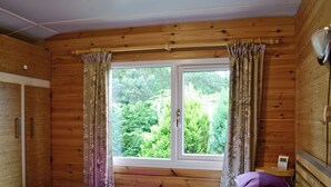A wooden cabin room with a bed, a window with curtains, and a view of greenery outside. - 2 bedroom accommodation in Nairn, Morayshire (Nairn, Morayshire)