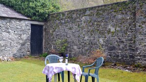 A small outdoor dining area with a table and two chairs, set against a stone wall and a building with a door.