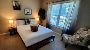 A bedroom with a bed, bedside lamps, a window with blinds, a chair, and wall art. - Horse Ranch Getaway (Richlands)