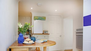 A small kitchen with a wooden table, a blue vase with flowers, a plate of biscuits, and a radio. - Westerlands Apartment (Torquay)