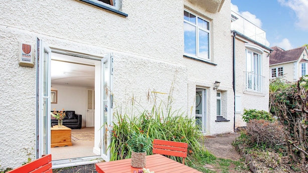 A white house with a patio table and chairs, a door leading to a living room, and a garden area with plants. - Westerlands Apartment (Torquay)