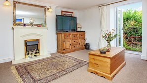 A living room with a fireplace, a television, a wooden cabinet, and a view of the garden through a sliding door. - Westerlands Apartment (Torquay)