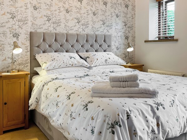 Een opgemaakt bed met bloemenbeddengoed, twee bedlampjes en een raam met zonwering.