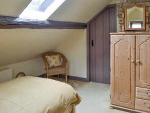 Ein Schlafzimmer mit schrägverdeckter Decke, einem Holzschrank, einem Korbstuhl und einem Bett mit beiger Bettdecke.