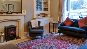 A cozy living room with a fireplace, a bookshelf, and a large window with curtains. - 4 bedroom accommodation in Coniston (Coniston)