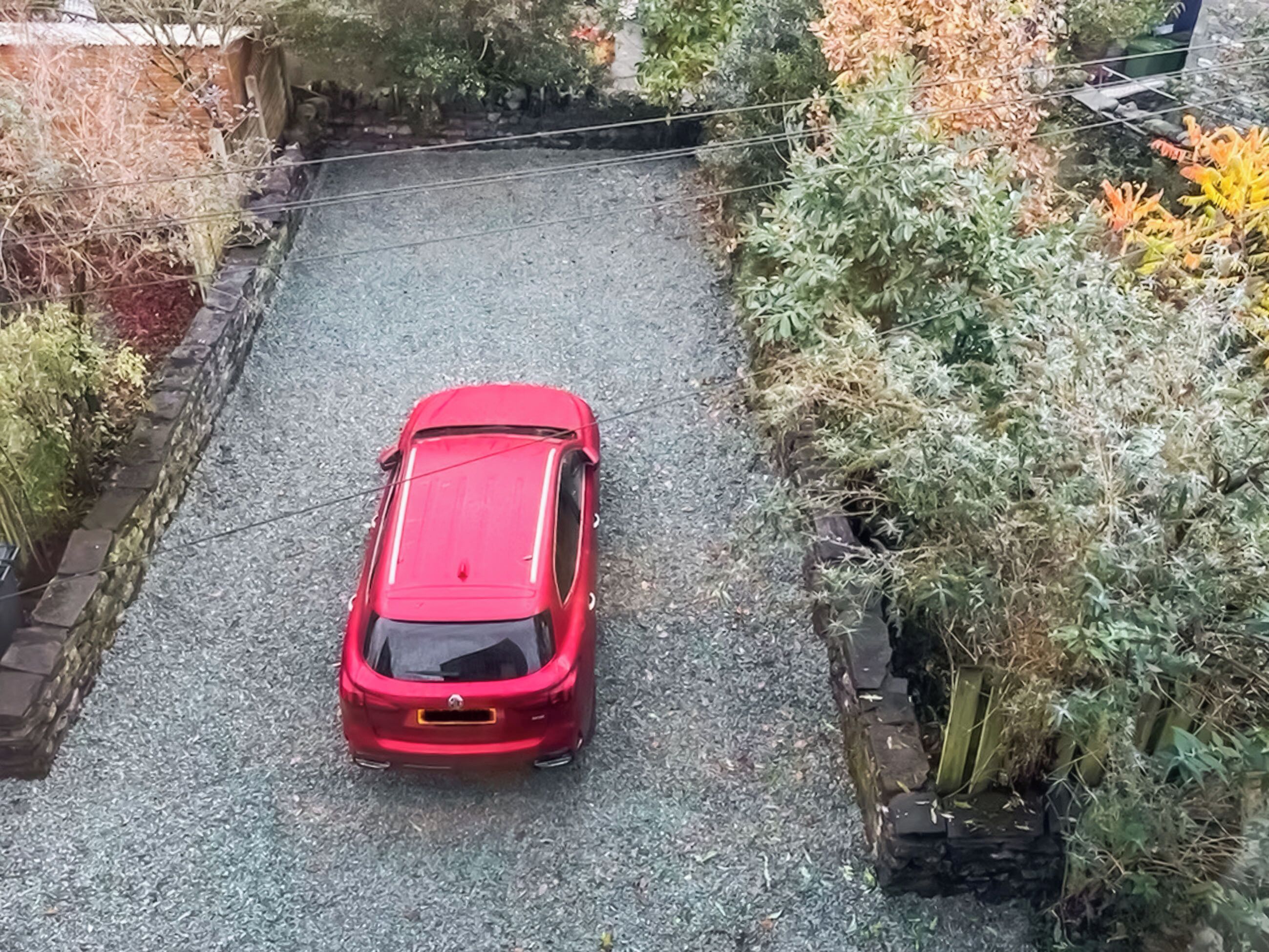 Une voiture rouge est garée sur une allée de gravier, entourée d’une végétation dense et d’un mur de briques.