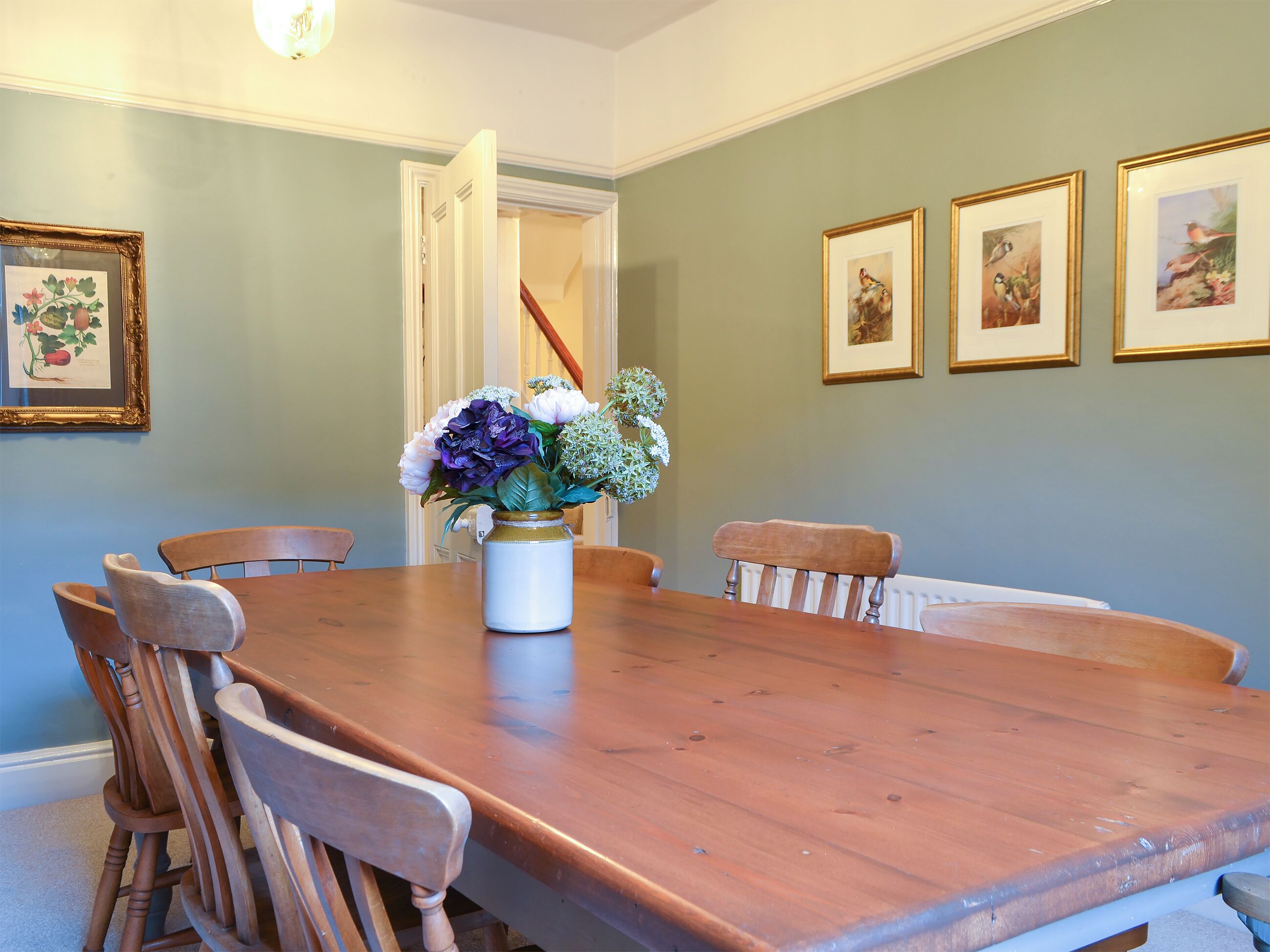 Une salle à manger avec une table et des chaises en bois, un vase rempli de fleurs et des tableaux encadrés accrochés au mur.