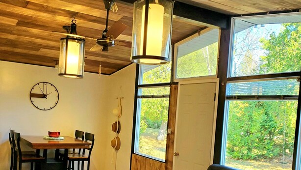 Ein modernes Wohnzimmer mit Holzboden, einem Essbereich mit Holztisch und -stühlen, einer schwarzen Ledersofa und großen Fenstern mit Blick auf Grünflächen.