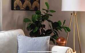 Una sala de estar con un sofá, un cojín a rayas, una planta en maceta, una lámpara de pie y una obra de arte en la pared.