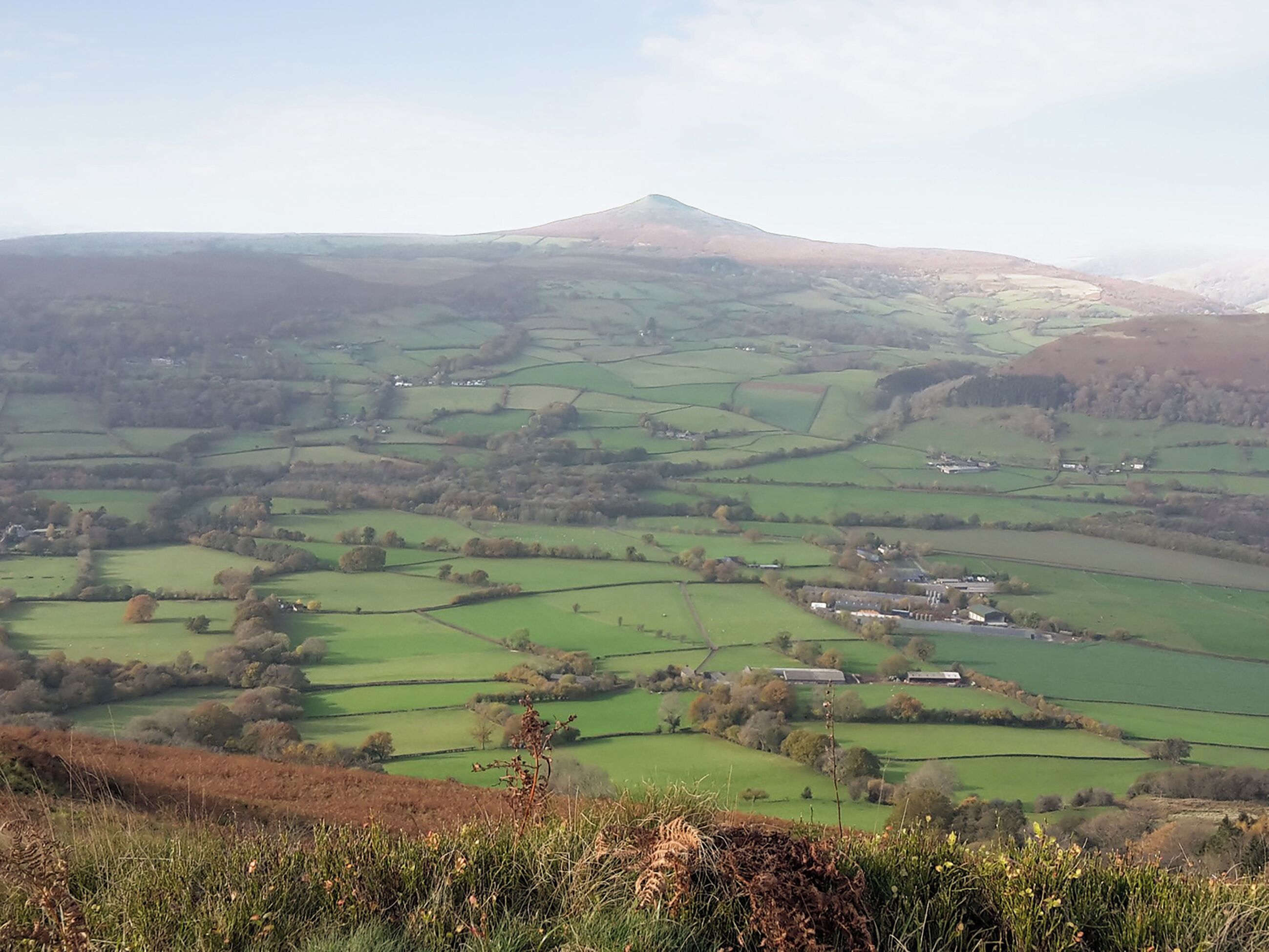 Une vue panoramique d’un paysage rural, avec de vastes champs verdoyants, des maisons éparpillées et une colline imposante à l’horizon.