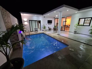 A modern outdoor pool area with a clear pool, a small waterfall feature, and a seating area with chairs and tables.