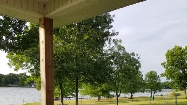 A covered patio with two Adirondack chairs and a small round table, overlooking a grassy area and a body of water. - Lake Front Gated Property  Private Dock 
Book 3 Nights - Get 4th Night Free.  (Wills Point)