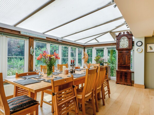 A conservatory with a dining table set for six, surrounded by wooden chairs.
