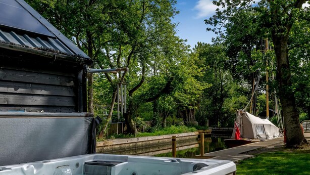 A hot tub in a wooded area with a wooden deck and a covered area in the background. - 5 bedroom accommodation in Ludham (Ludham)
