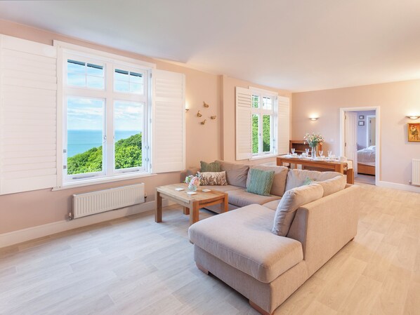 A spacious living room with a large sofa, a dining table, and a view of the sea through large windows. - 2 bedroom accommodation in Woody Bay, Exmoor (Woody Bay, Exmoor)
