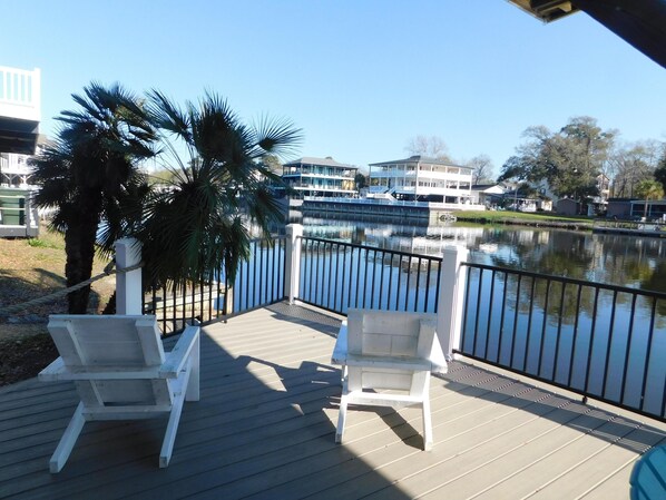 A deck with two white chairs, a palm tree, and a view of a lake with buildings in the background. - Ocean Lakes 308 | Fish off the deck, relax and enjoy the salt air breeze (Myrtle Beach)