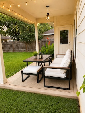 A covered patio with a table and two sofas, a string of lights, and a wooden fence in the background. - Modern South Austin Home|King Bed • Gym • Patio • Sleeps 6- Ideal for long stays (Austin)