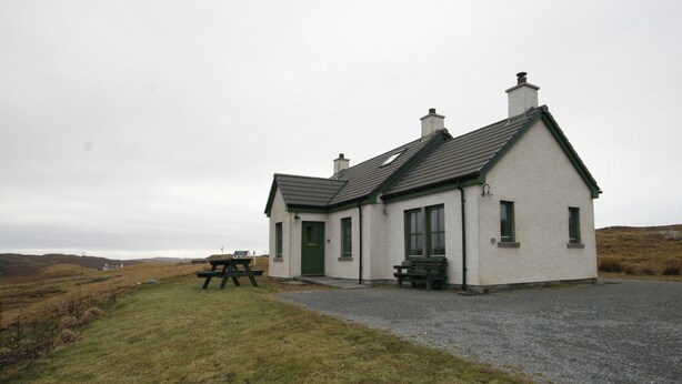 A small, single-story house with a green door and windows, a gravel driveway, and a picnic table in the foreground.