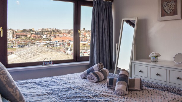 A bedroom with a large window, a bed with patterned bedding, a dresser with a mirror, and a view of a marina and buildings. - Situated in Whitby’s old town. (Whitby)