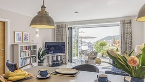 A modern living room with a TV, bookshelves, a dining area with fruit and cups, and a view of the outdoors. - 1 bedroom accommodation in 3 Coombe Close (3 Coombe Close)