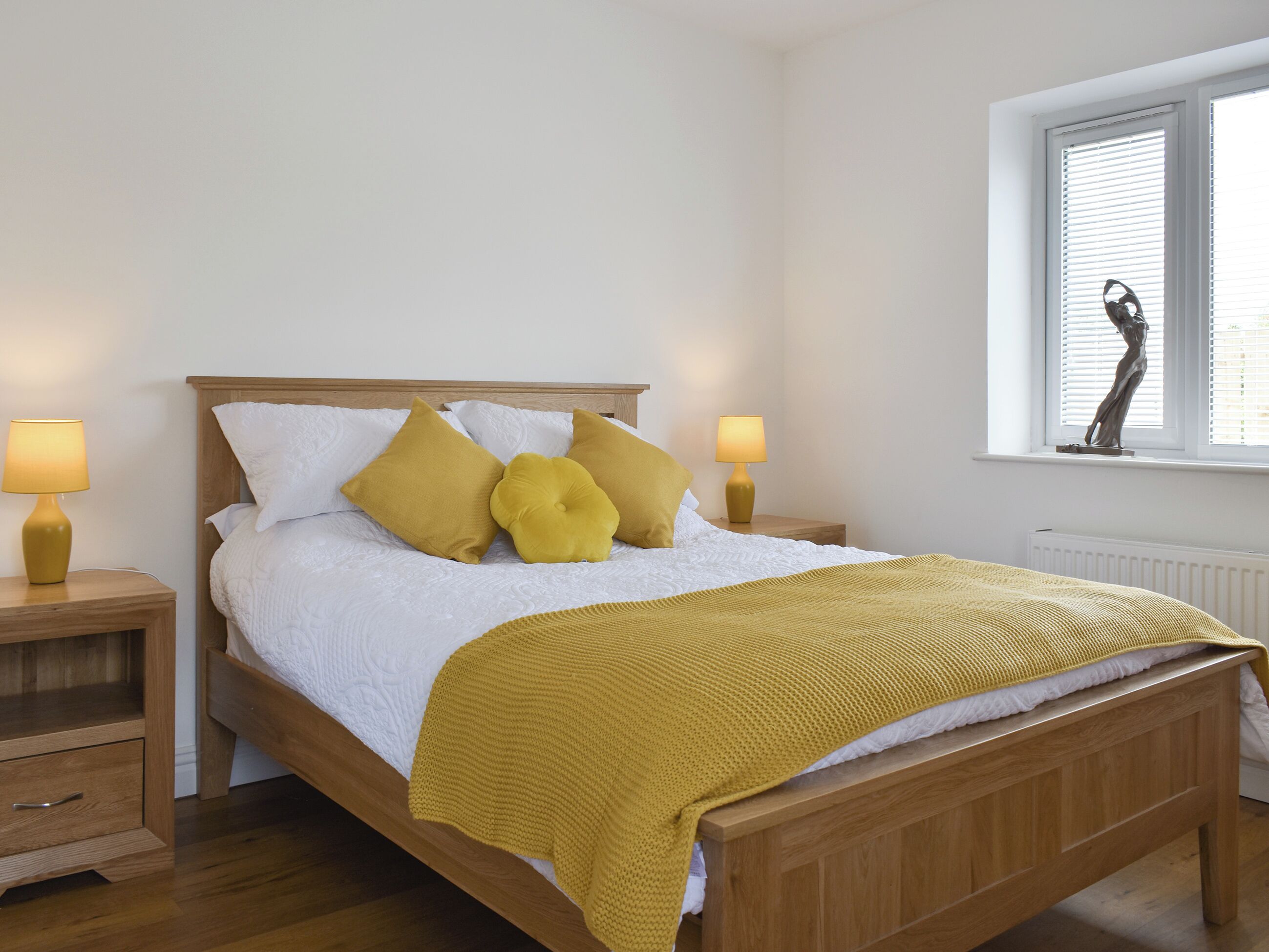 Ein ordentlich bezogenes Bett mit Holzbettgestell, zwei Nachttische mit Lampen, ein Fenster mit Jalousien und eine Heizung.