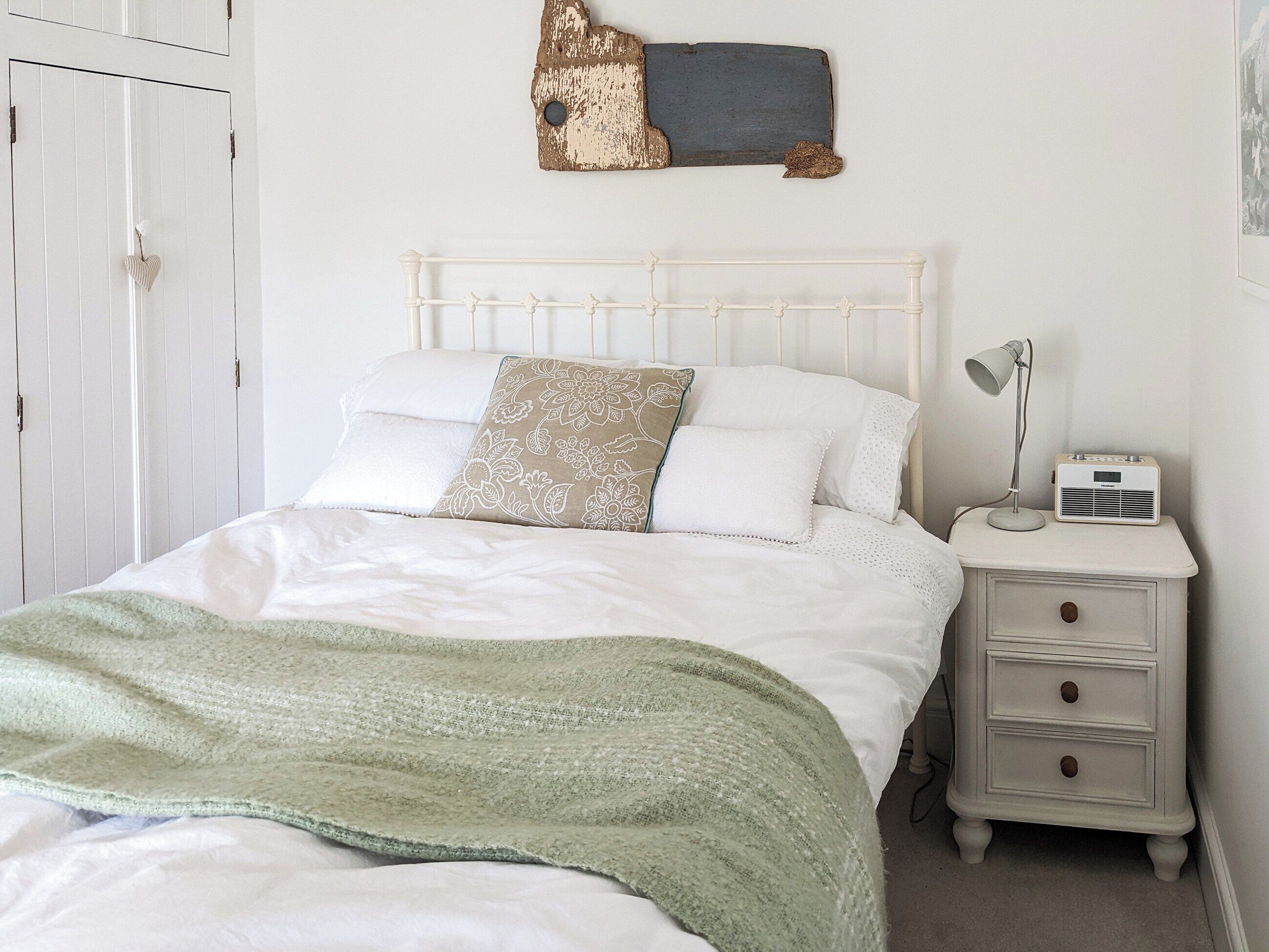 A neatly made bed with a green blanket, a bedside lamp, and a small white nightstand with drawers.