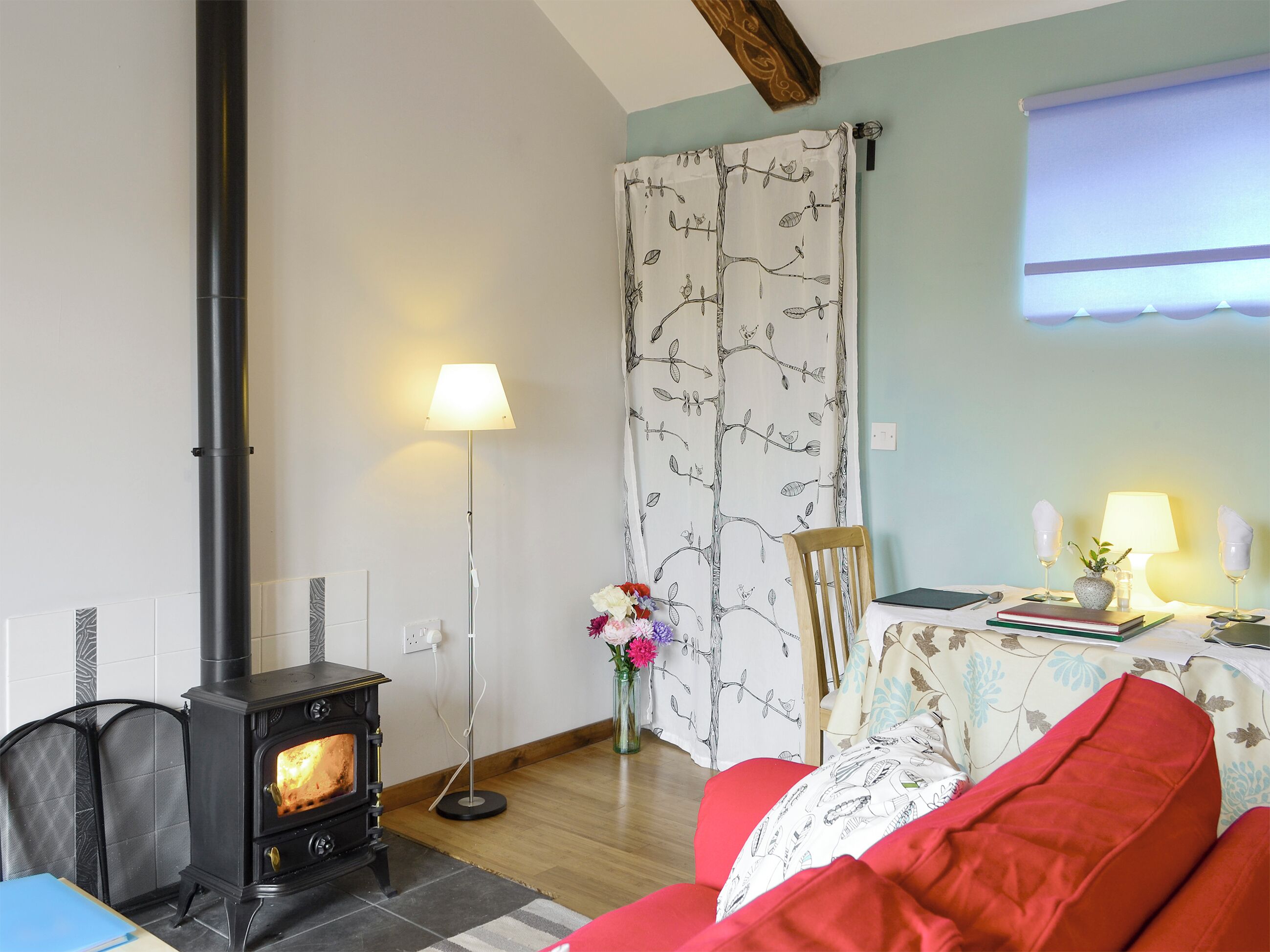 A cozy living room with a wood-burning stove, a red sofa, a table with a lamp, and a window with a view.