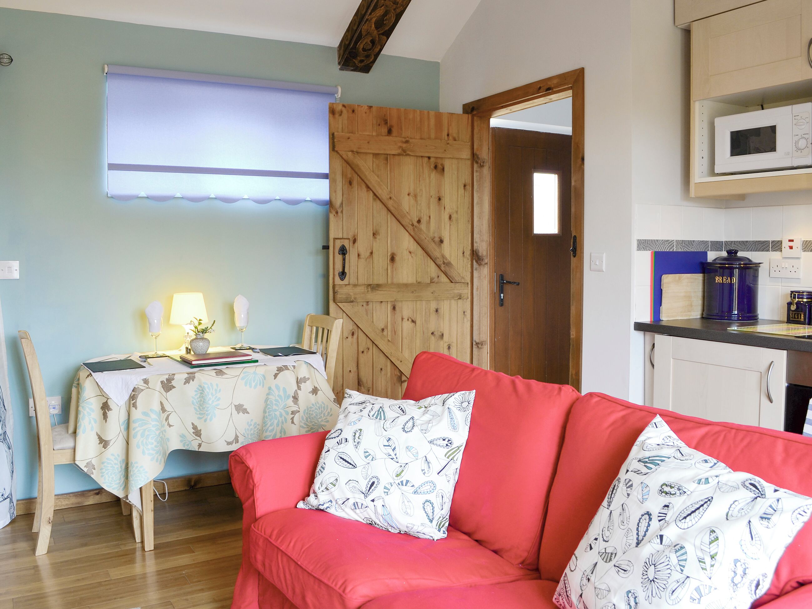 A cozy room with a wooden door, a dining table with a floral tablecloth, a red sofa with patterned cushions, and a kitchen area with a microwave and bread container.