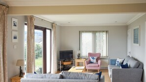 A living room with a grey sofa, a pink armchair, and a grey armchair. There is a TV on a stand, a small table with a lamp, and a window with blinds.
