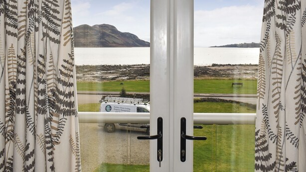 A view through a window with curtains, featuring a vehicle on a road and a body of water with mountains in the background.