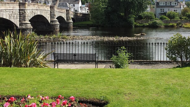 A stone bridge over a river, a bench, and a grassy area with pink flowers.