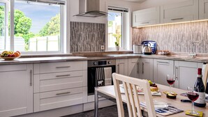 A modern kitchen with white cabinets, a built-in oven, a dining table with chairs, and a window with a view of greenery.
