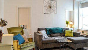 A living room with a green armchair, a grey sofa with a green throw, a small table, and a large abstract wall art piece.