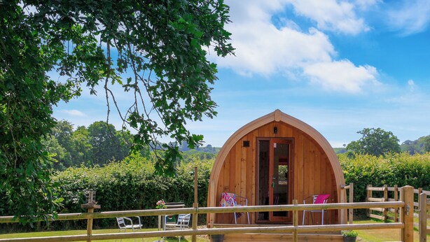 Una baita di legno con tetto spiovente, circondata da una recinzione in legno e vegetazione lussureggiante.
